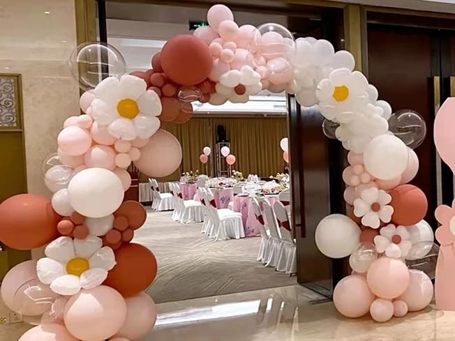 Arc de baloane crem, roz și maro cu flori albe mari, montat la intrarea într-un salon.